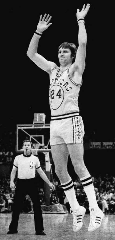 Rick Barry jumping while a referee stands behind him.